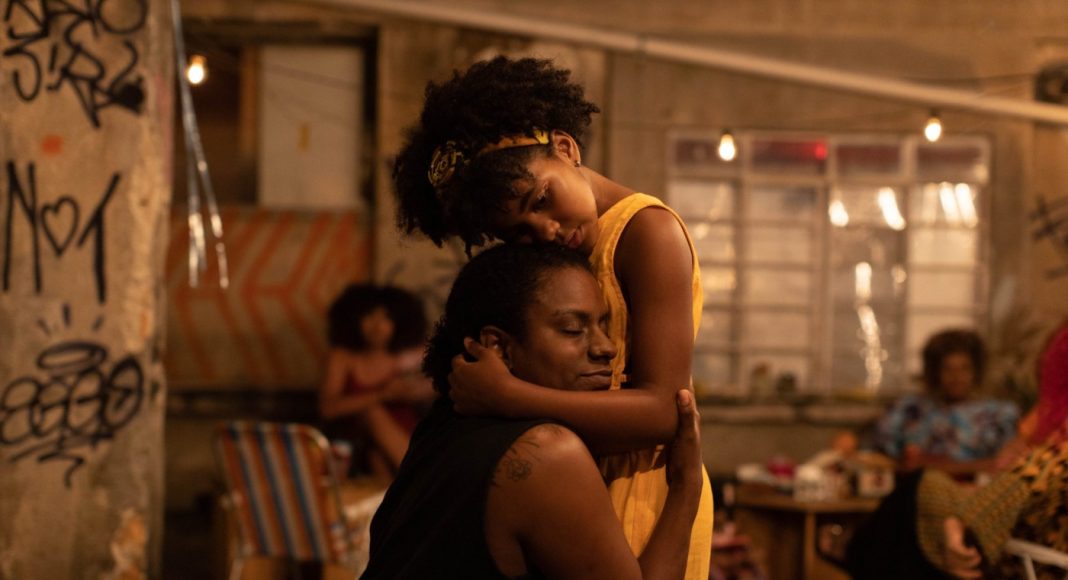 mae Two people share a comforting hug in a dim, graffiti-filled indoor space with warm lights in the background.