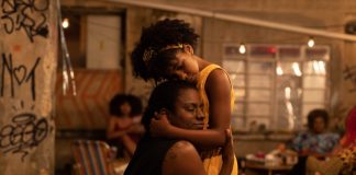 Two people share a comforting hug in a dim, graffiti-filled indoor space with warm lights in the background.