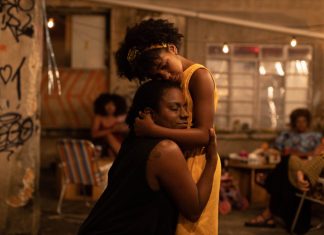 Two people share a comforting hug in a dim, graffiti-filled indoor space with warm lights in the background.