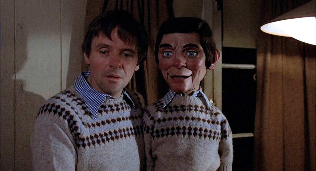magia negra (1) Two people stand close together in a dim room, wearing matching knotted-pattern sweaters, with the right figure a cracked-faced ventriloquist dummy.