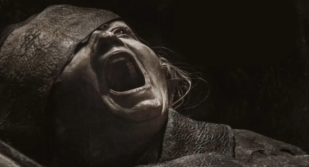 maldição da múmia Person screaming with mouth wide open, wearing a tattered hood and rough clothing in a dark scene.