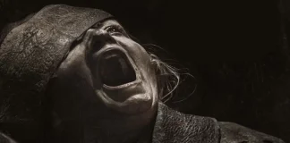 Person screaming with mouth wide open, wearing a tattered hood and rough clothing in a dark scene.