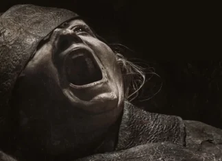 Person screaming with mouth wide open, wearing a tattered hood and rough clothing in a dark scene.