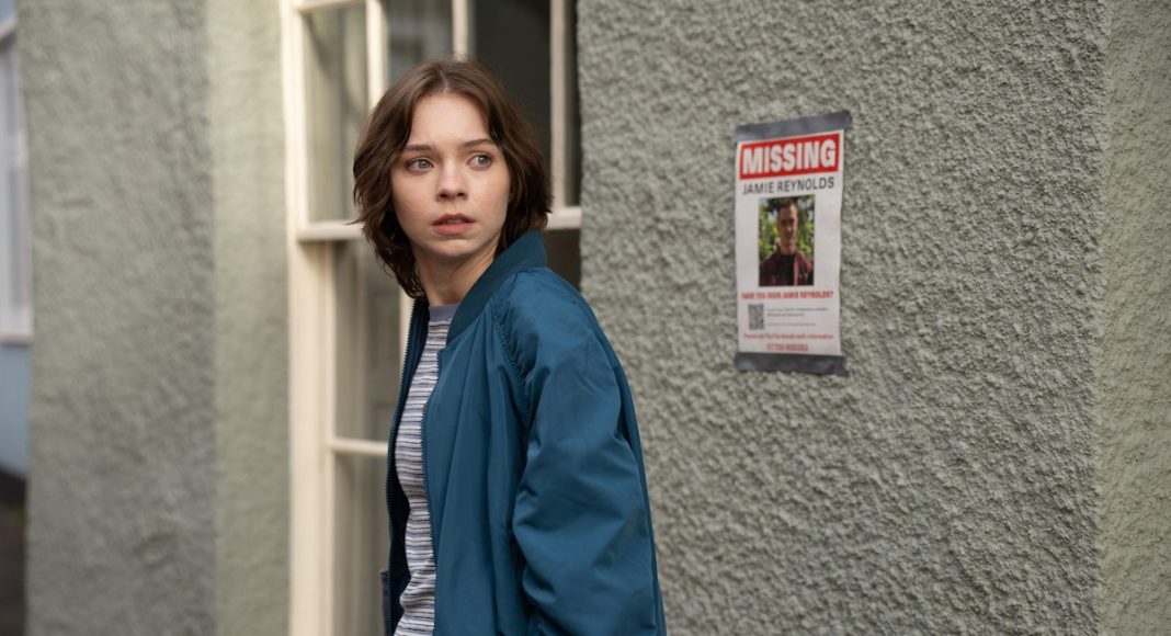 Young woman in a blue jacket glances to her left beside a textured wall with a missing person poster nearby.
