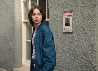 Young woman in a blue jacket glances to her left beside a textured wall with a missing person poster nearby.
