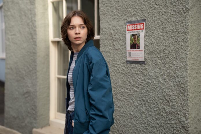 Young woman in a blue jacket glances to her left beside a textured wall with a missing person poster nearby.