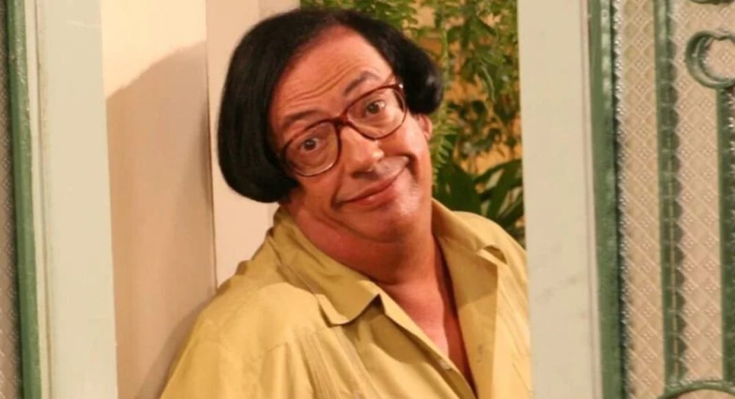 marcos oliveira-a grande familia (1) Man with red-framed glasses and a yellow shirt leaning against a doorway, smiling at the camera.