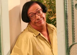 Man with red-framed glasses and a yellow shirt leaning against a doorway, smiling at the camera.