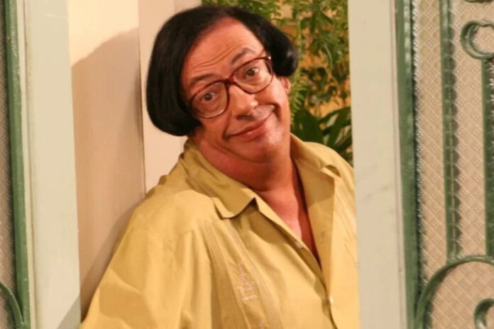 Man with red-framed glasses and a yellow shirt leaning against a doorway, smiling at the camera.