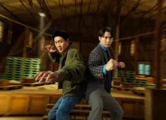 Two men in a warehouse strike back-to-back fighting stances; one in a green jacket extends a punch.