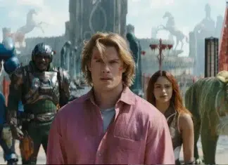 ‘Mestres do Universo’: Nicholas Galitzine detalha preparação física para viver o Príncipe Adam; Confira! Film still: a man in a pink shirt leads a diverse group through a bustling fantasy city with a tiger nearby. (Note: concise summary focusing on the scene and characters.)