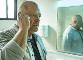 Bald man in a gray shirt talking on a cellphone in a bathroom, his reflection in the mirror showing him touching his head with a bruise visible near the temple.