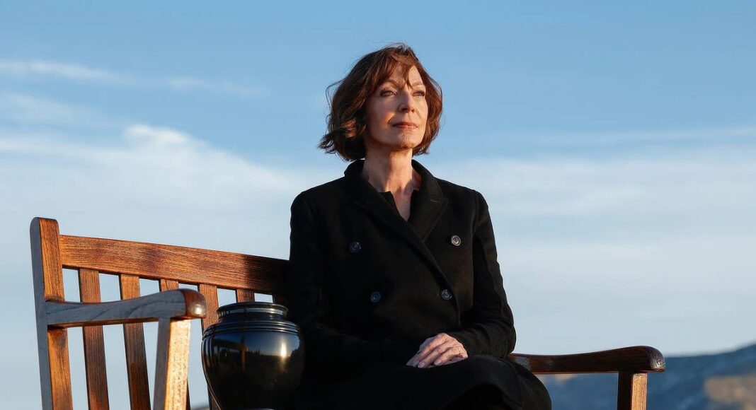 Contemplative woman sits on a wooden bench outdoors, with a black urn beside her against a blue sky and distant hills.