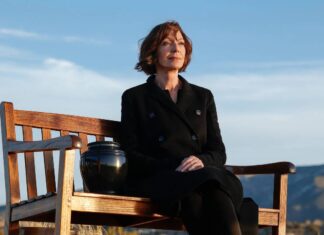 Allison Janney vive uma viúva enlutada no trailer de ‘Miss You, Love You’, novo DRAMA da HBO Contemplative woman sits on a wooden bench outdoors, with a black urn beside her against a blue sky and distant hills.