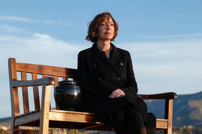 Contemplative woman sits on a wooden bench outdoors, with a black urn beside her against a blue sky and distant hills.