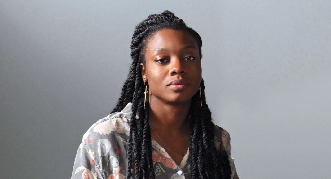 Portrait of a woman with long box braids, wearing a floral gray shirt, looking calmly at the camera against a gray backdrop.