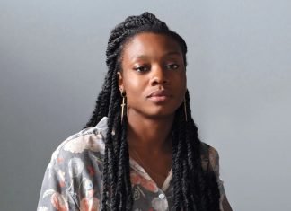 Portrait of a woman with long box braids, wearing a floral gray shirt, looking calmly at the camera against a gray backdrop.