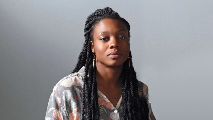 Portrait of a woman with long box braids, wearing a floral gray shirt, looking calmly at the camera against a gray backdrop.