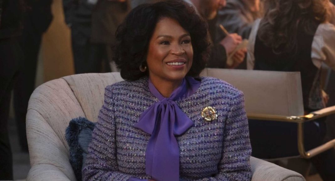 Smiling woman in a purple bow-tied blouse and tweed blazer sits on a beige armchair at a social event.