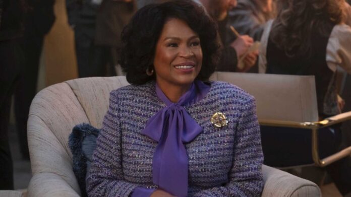 Smiling woman in a purple bow-tied blouse and tweed blazer sits on a beige armchair at a social event.