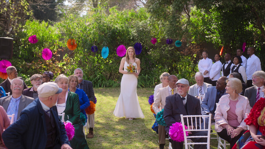 Bride in a white gown holding a bouquet walks down the aisle at an outdoor garden wedding ceremony.