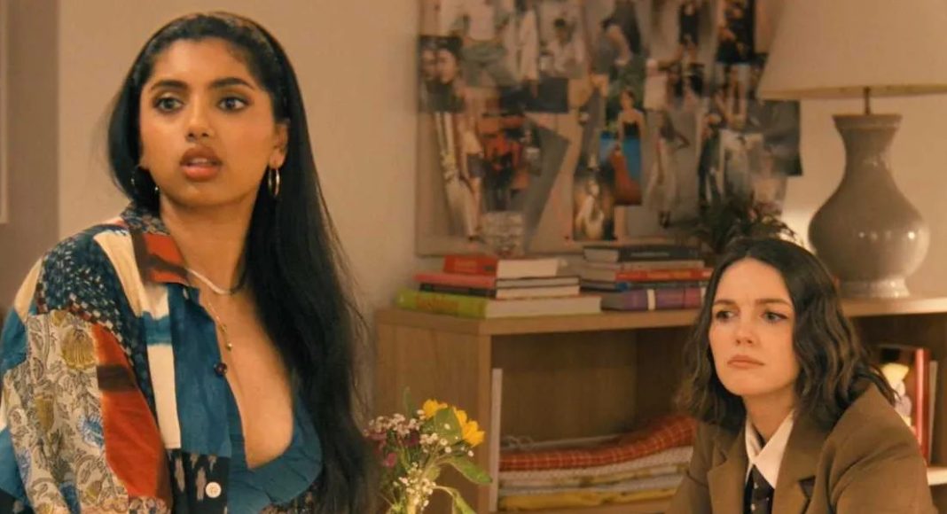 Two women in a living room: one with long dark hair in a colorful patchwork shirt speaks, the other in a brown blazer listens nearby.