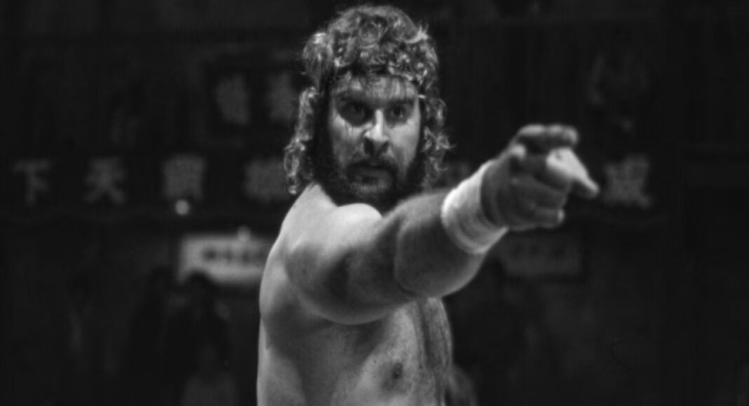 Bearded, shirtless wrestler with long hair points toward the camera in a black-and-white scene.