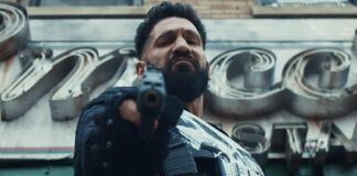 A rugged man in tactical gear points a pistol at the camera, in a gritty urban setting with metallic signage behind him.