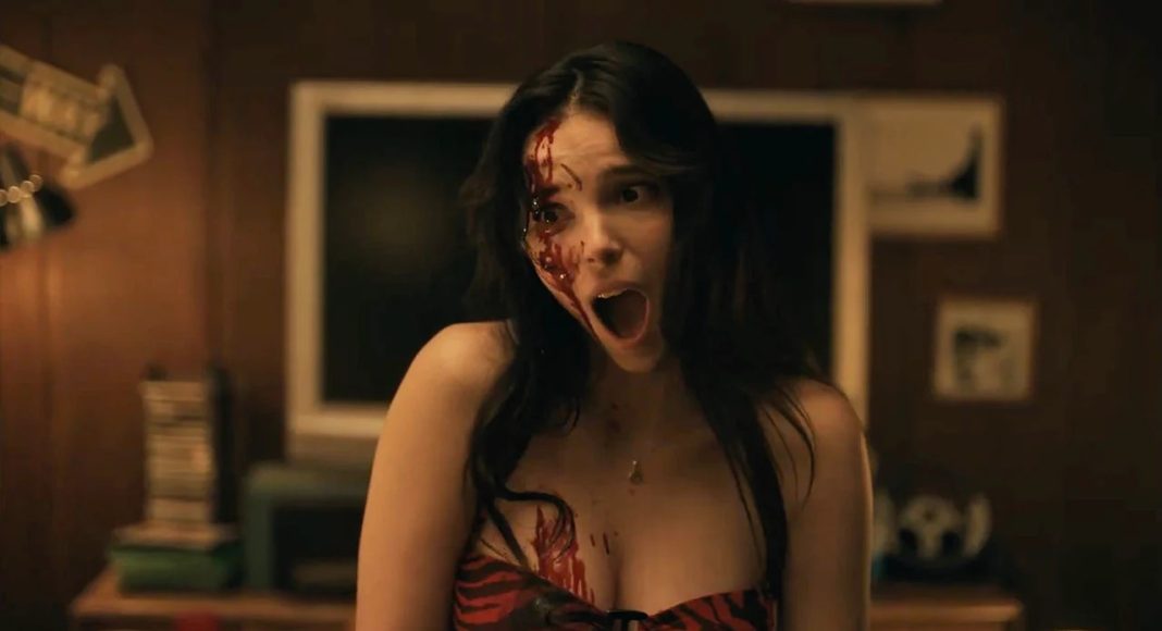 Woman with blood on her face and chest, mouth open in a scream in a dimly lit room.