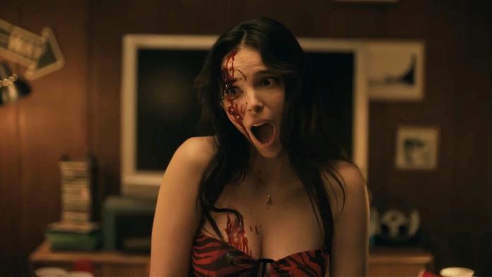 Woman with blood on her face and chest, mouth open in a scream in a dimly lit room.