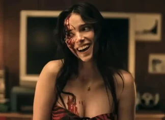 Woman with black hair, blood smeared on the left side of her face and chest, smiling in a dimly lit room.