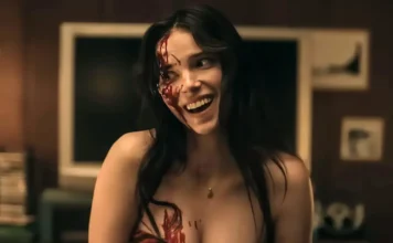 Woman with black hair, blood smeared on the left side of her face and chest, smiling in a dimly lit room.
