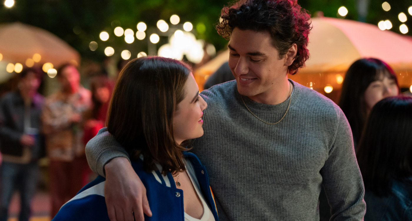 Couple with the man’s arm around the woman, smiling at each other at an outdoor evening gathering with string lights in the background.