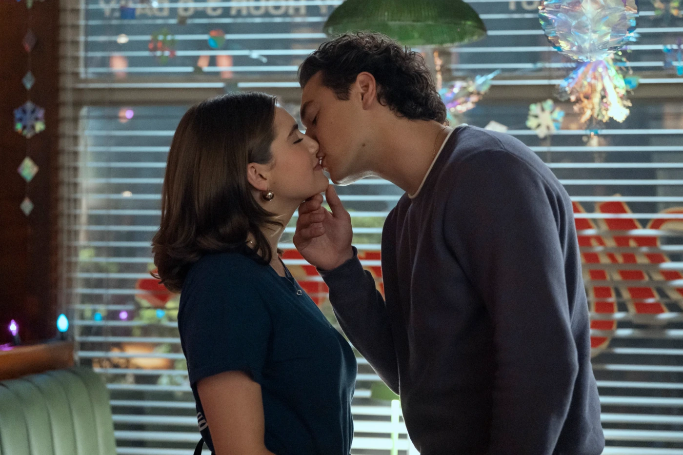 A couple kissing in a cozy indoor setting, with one hand gently cupping the other’s chin as they lean in. behind them are striped blinds and festive decorations.