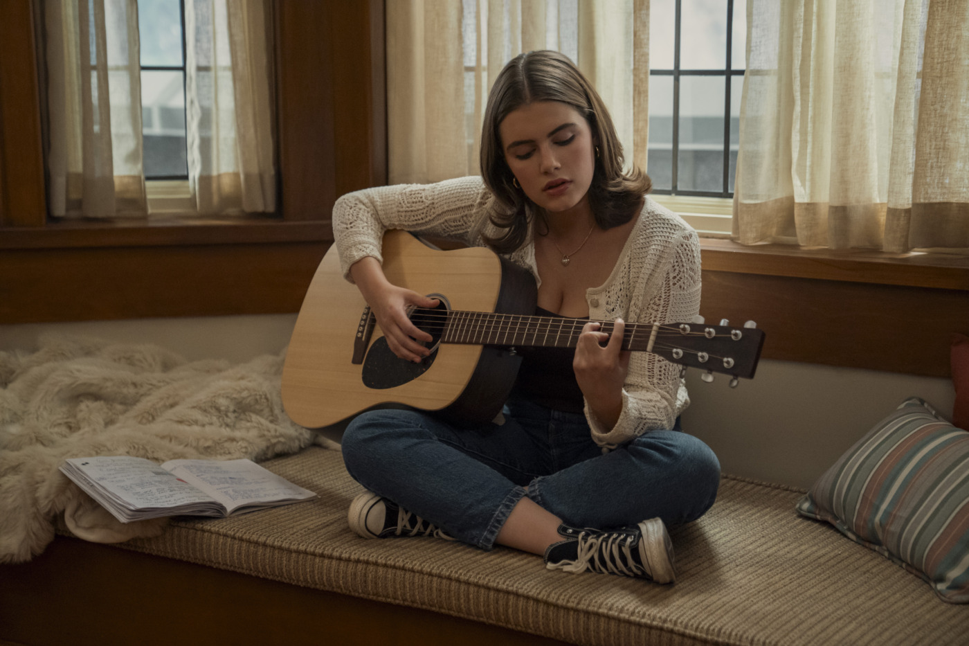Young woman sits cross-legged on a cushioned bench, strumming an acoustic guitar with an open notebook nearby.