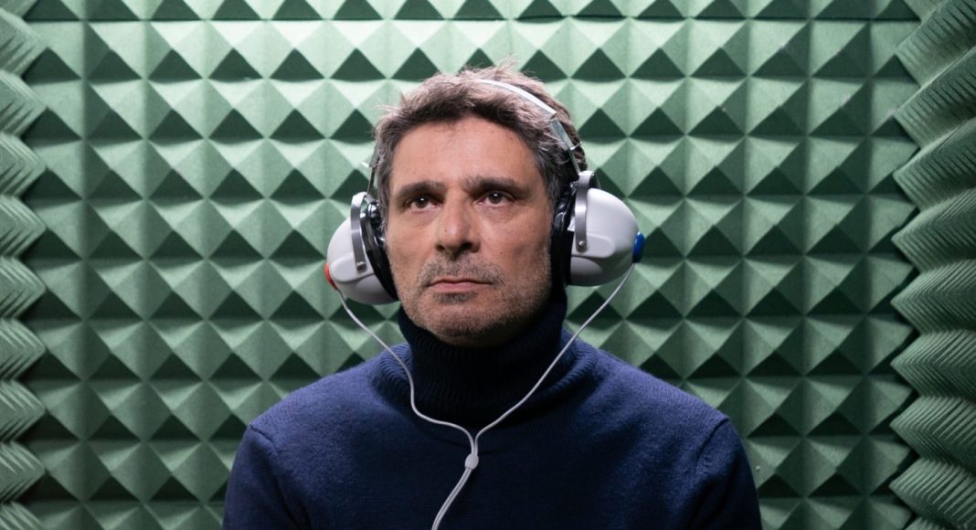 Man wearing a dark blue turtleneck with over-ear headphones in a green acoustic foam chamber.
