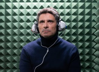 Man wearing a dark blue turtleneck with over-ear headphones in a green acoustic foam chamber.