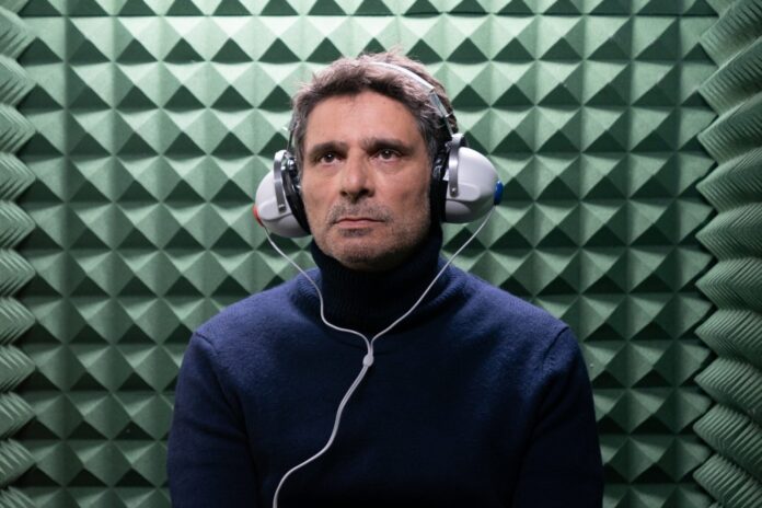Man wearing a dark blue turtleneck with over-ear headphones in a green acoustic foam chamber.