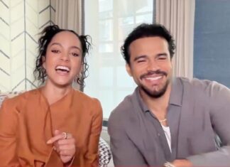 Two people sit on a striped couch, smiling and laughing at the camera in a bright room with patterned wallpaper behind them.