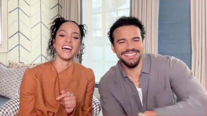 Two people sit on a striped couch, smiling and laughing at the camera in a bright room with patterned wallpaper behind them.
