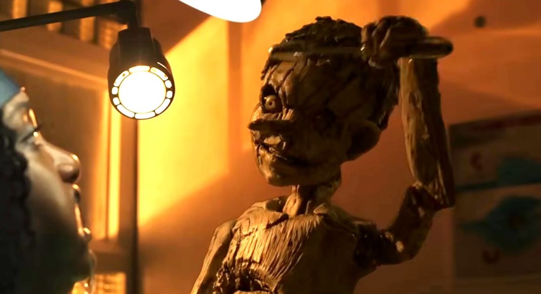 Stop-motion wooden puppet raises its arm under a warm desk lamp while a person watches in a dim workshop.
