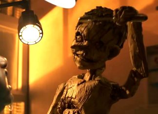 Stop-motion wooden puppet raises its arm under a warm desk lamp while a person watches in a dim workshop.