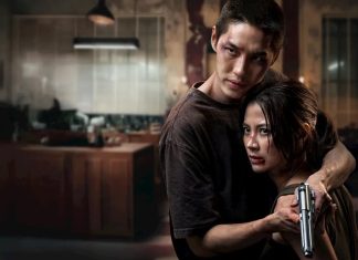 Man and woman in a dim kitchen; the man holds the woman, who grips a handgun and looks wary or scared.