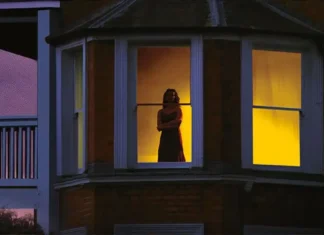‘Remain’: M. Night Shyamalan afirma que seu novo thriller teve a MAIOR média de aprovação de sua carreira Woman standing in a lit upstairs window, arms crossed, warm yellow light inside the room