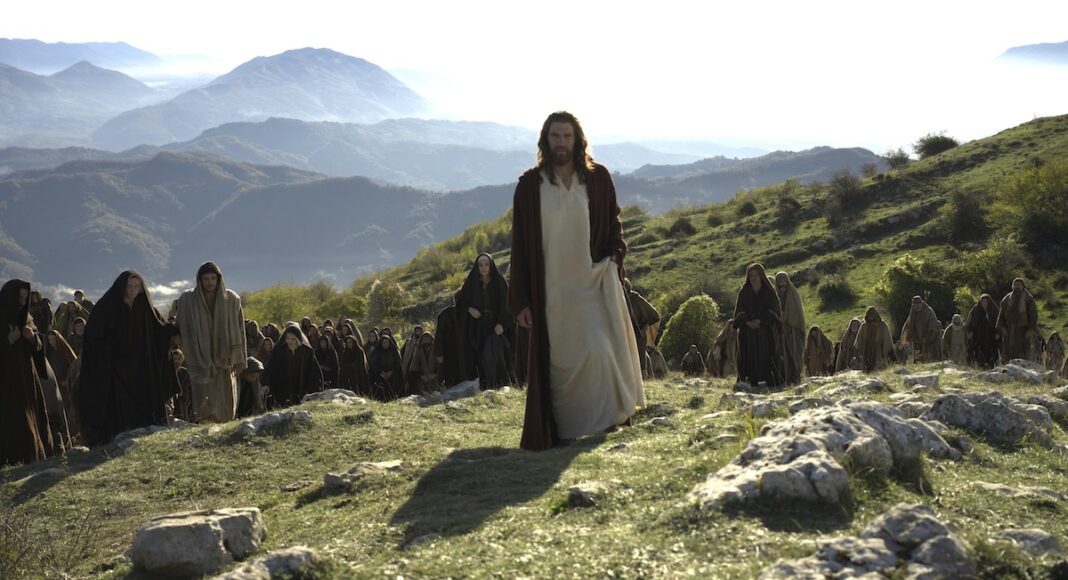 Man in white robes and red cloak leads a crowd of robed followers across a sunlit hillside with distant mountains.