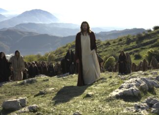Man in white robes and red cloak leads a crowd of robed followers across a sunlit hillside with distant mountains.