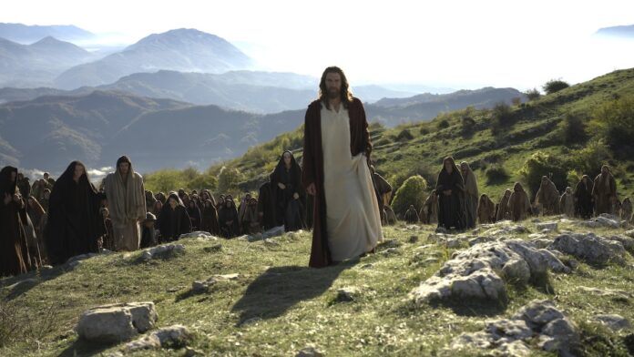 Man in white robes and red cloak leads a crowd of robed followers across a sunlit hillside with distant mountains.