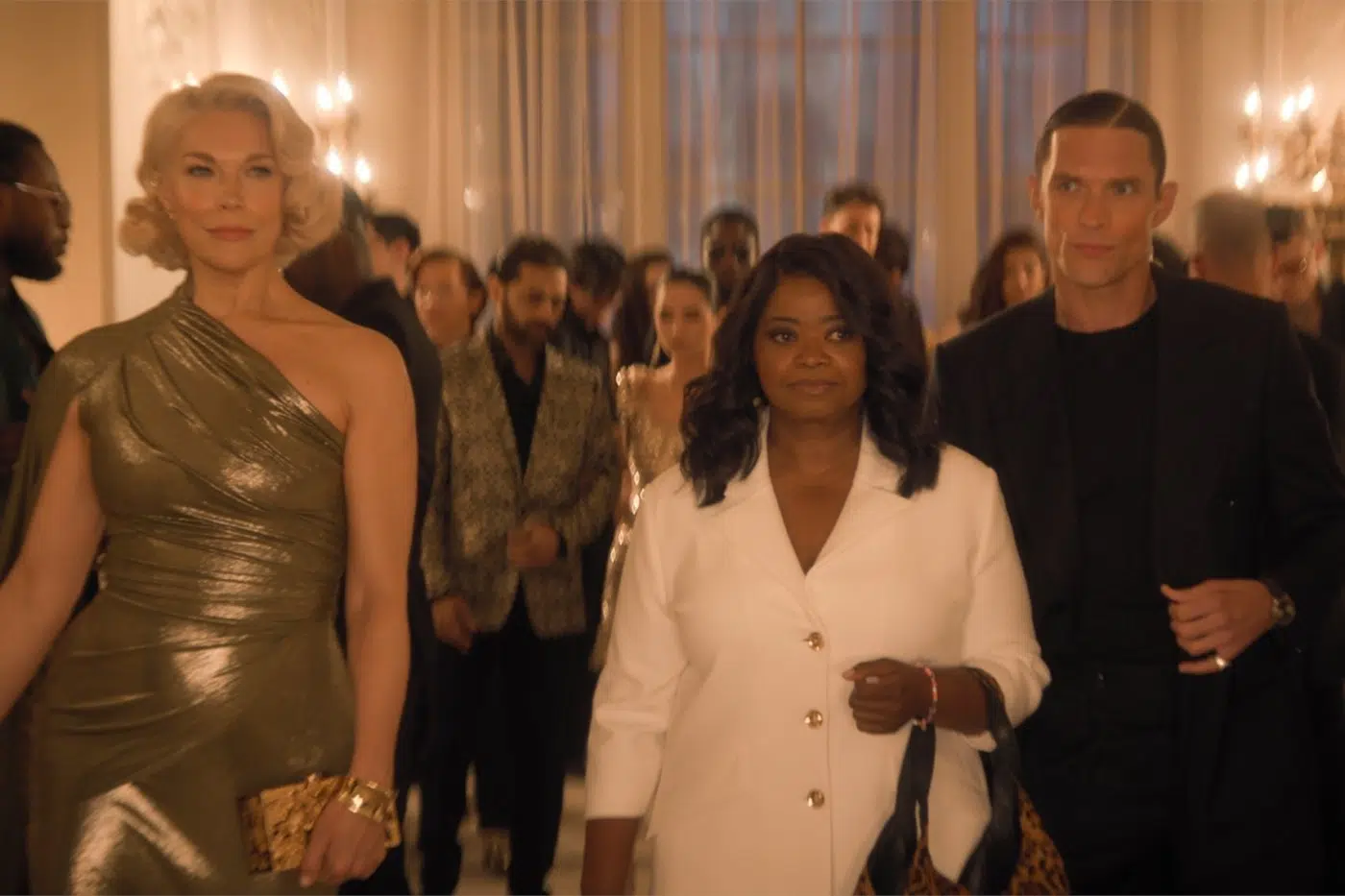 Three people lead a formal party crowd: a blonde in a gold one-shoulder dress on the left, a woman in a white blazer center, and a man in black on the right.