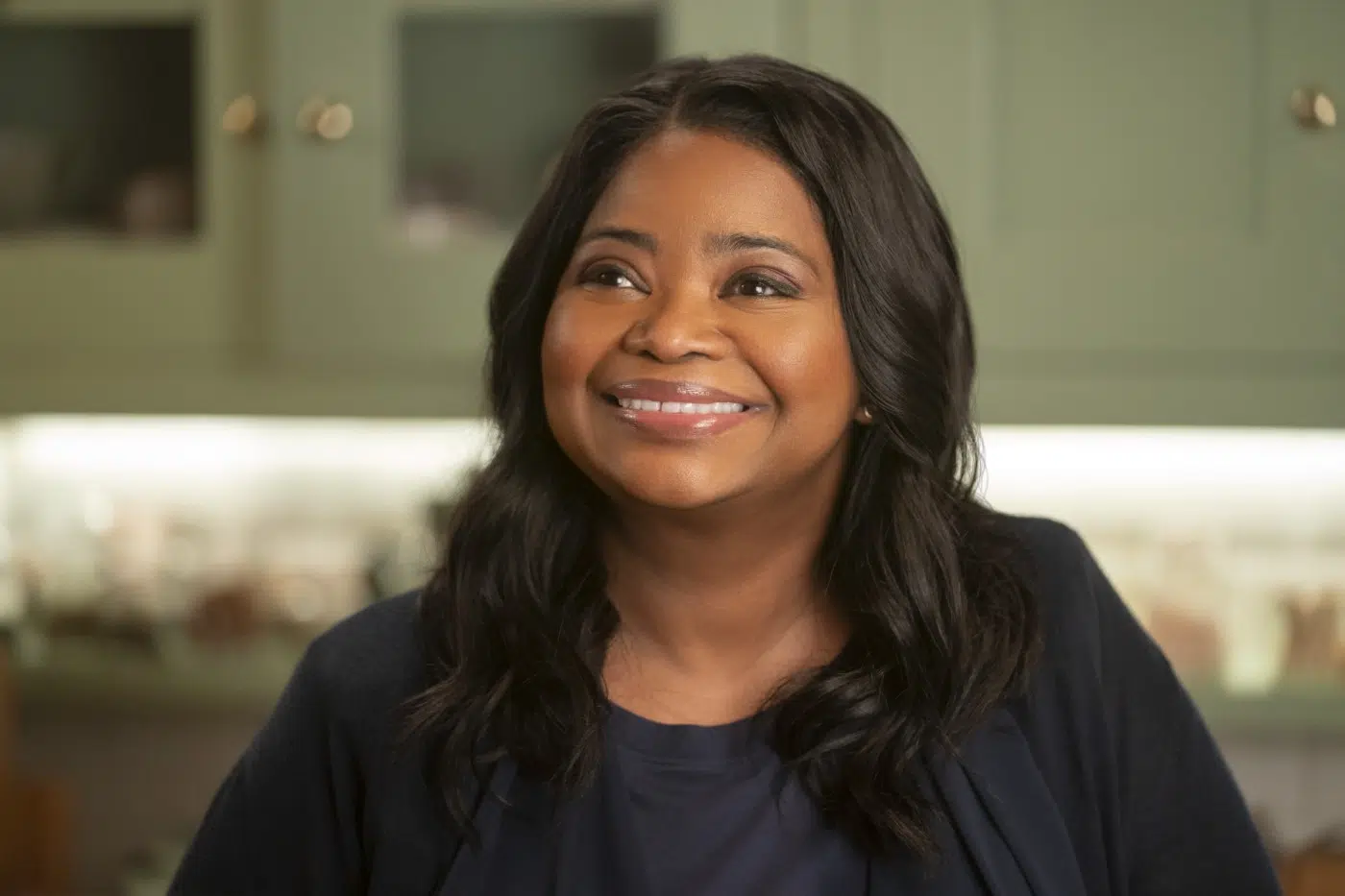 Smiling woman with dark hair in a navy top, portrait-style photo indoors with a blurred green kitchen background.
