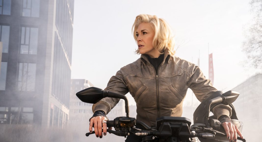 ride or die 3 Woman in a brown jacket sits on a black motorcycle in an urban area, looking to the side with a helmet resting on the right handlebar.